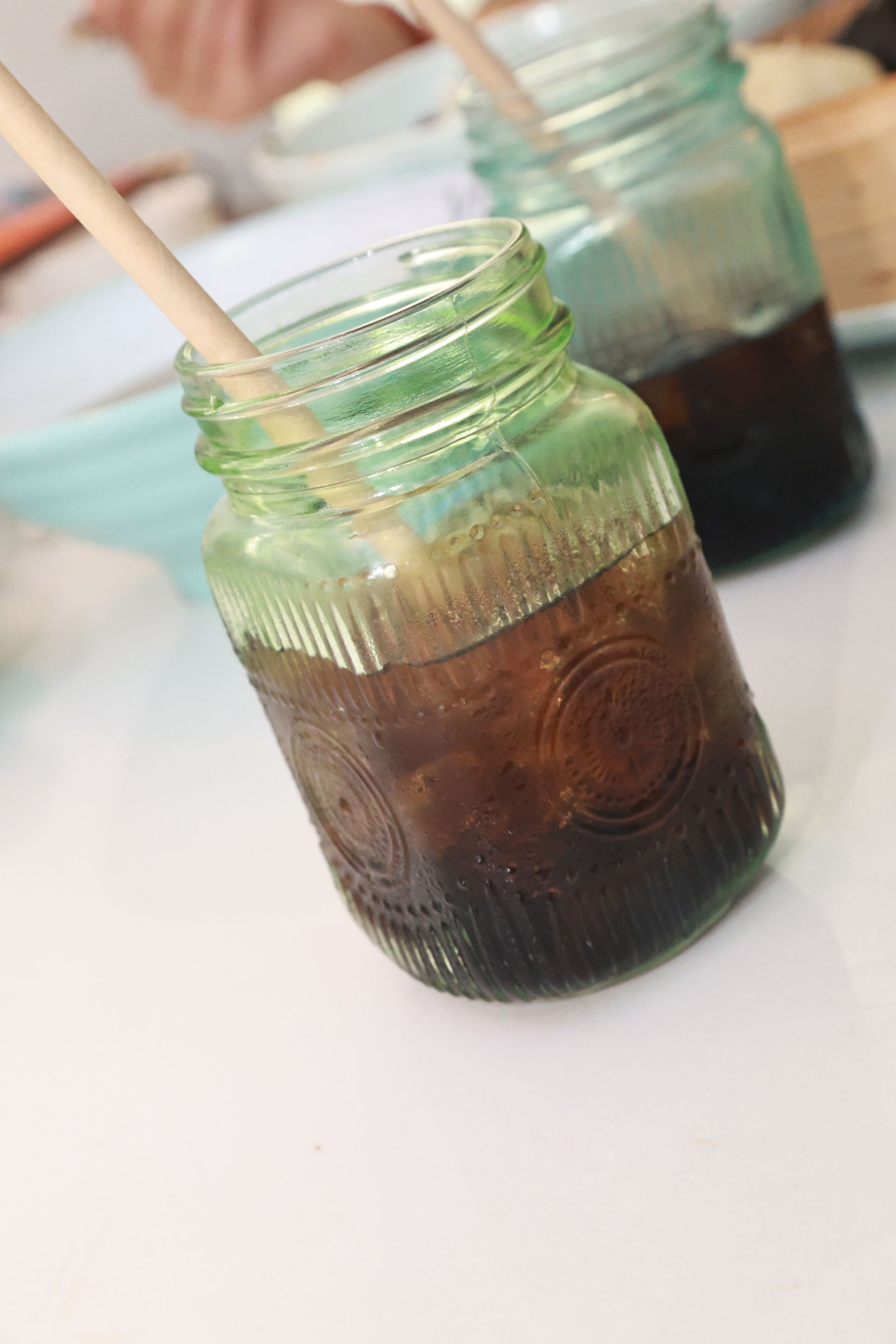 Zwei Gläser mit dunklem Getränk und Holzrührstäbchen auf einem weißen Tisch