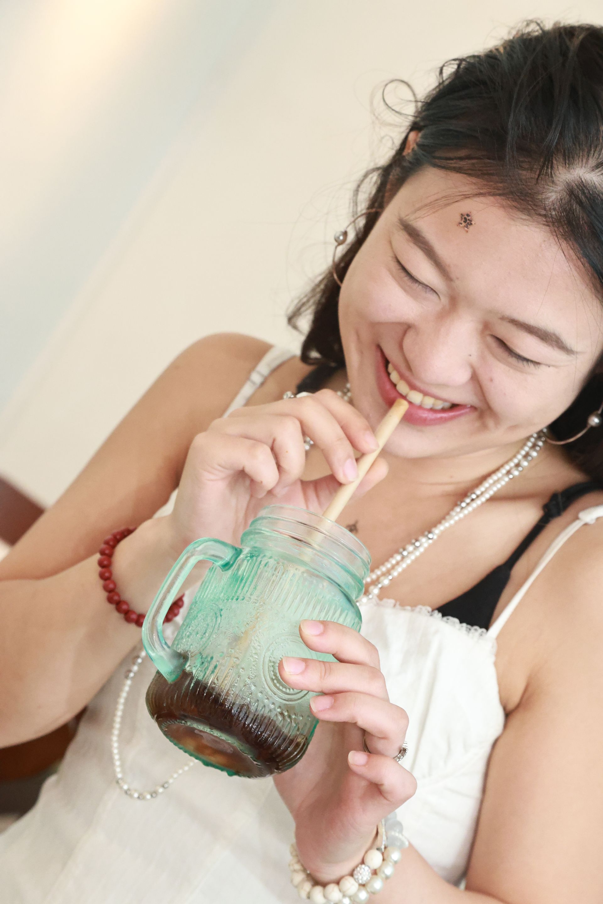 Lächelnde Person, die durch einen Strohhalm aus einem Glas mit Schraubverschluss einen dunklen Drink trinkt und ein weißes Oberteil trägt.