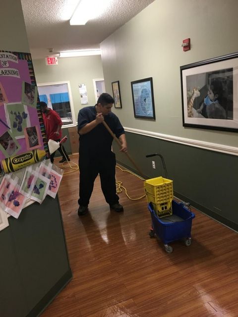 Man mopping a wet hallway floor with a mop and bucket. Hallway has artwork and a bulletin board.