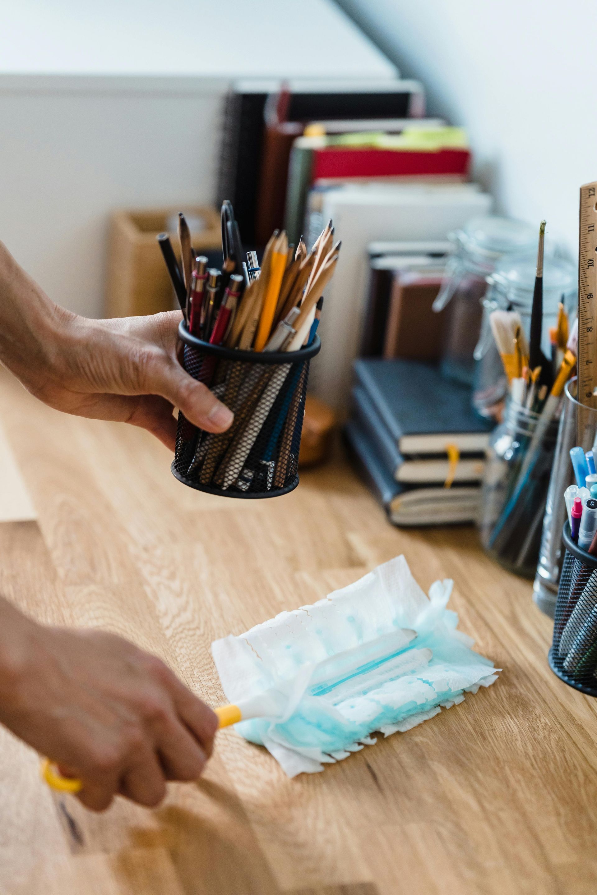 A person dusting a wooden desk with a hand duster, pencils in a holder nearby.