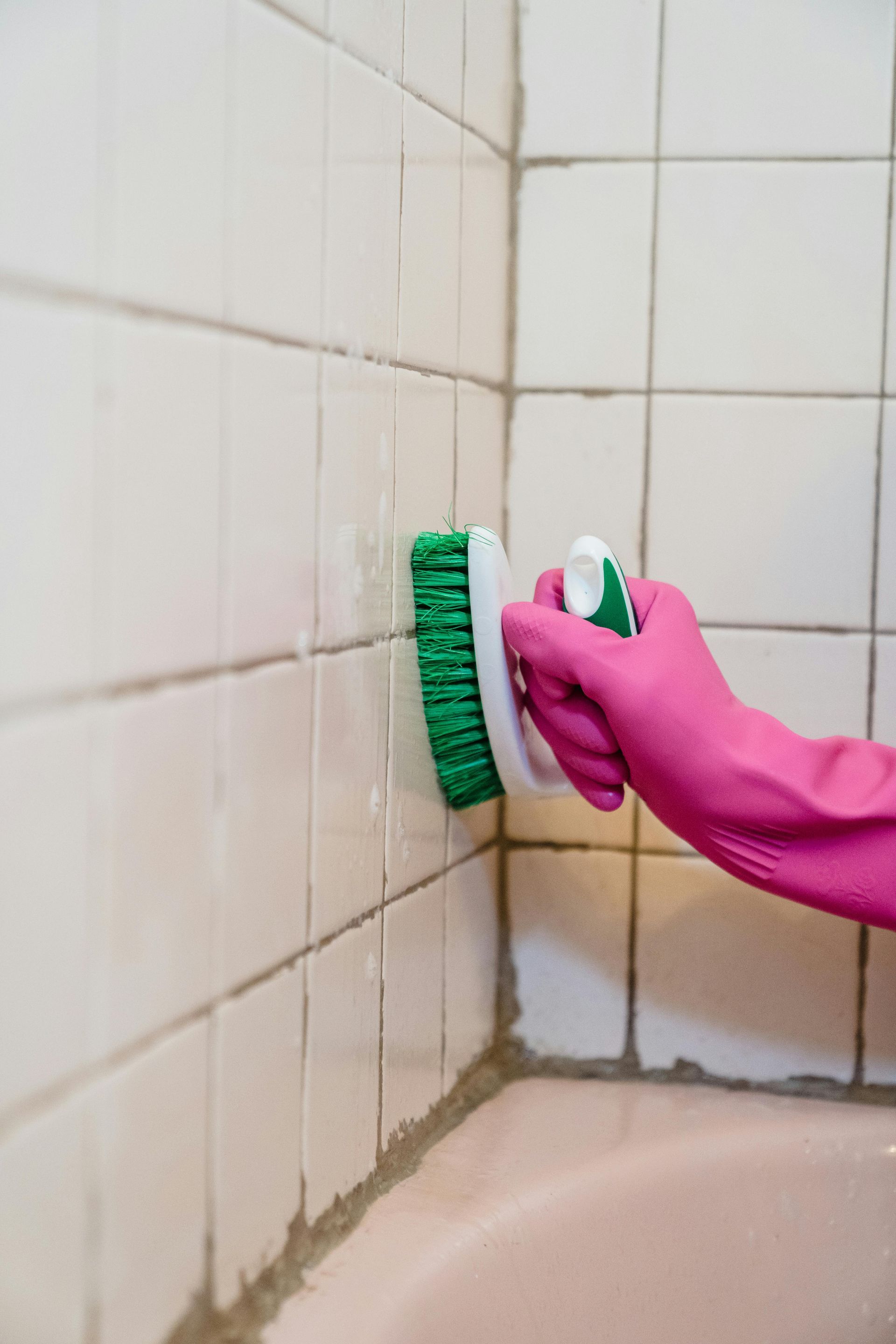 Person wearing pink gloves scrubbing bathroom tiles with a green brush.