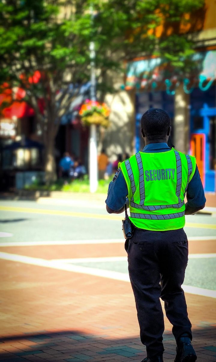 A security professional wearing a bright neon high-visibility vest and dark uniform walking through a sunny city plaza.