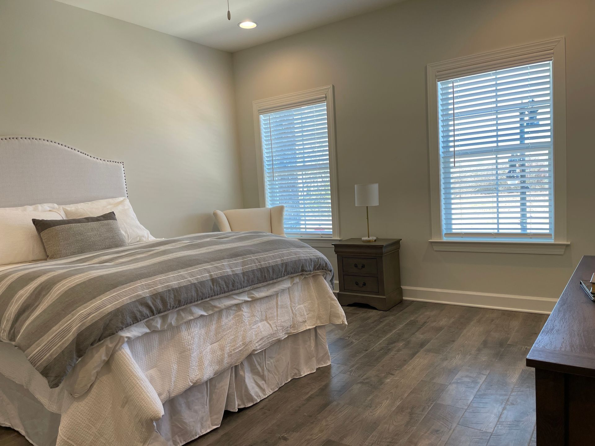 Bedroom with bed, nightstand, two windows with blinds, and wood-look flooring.