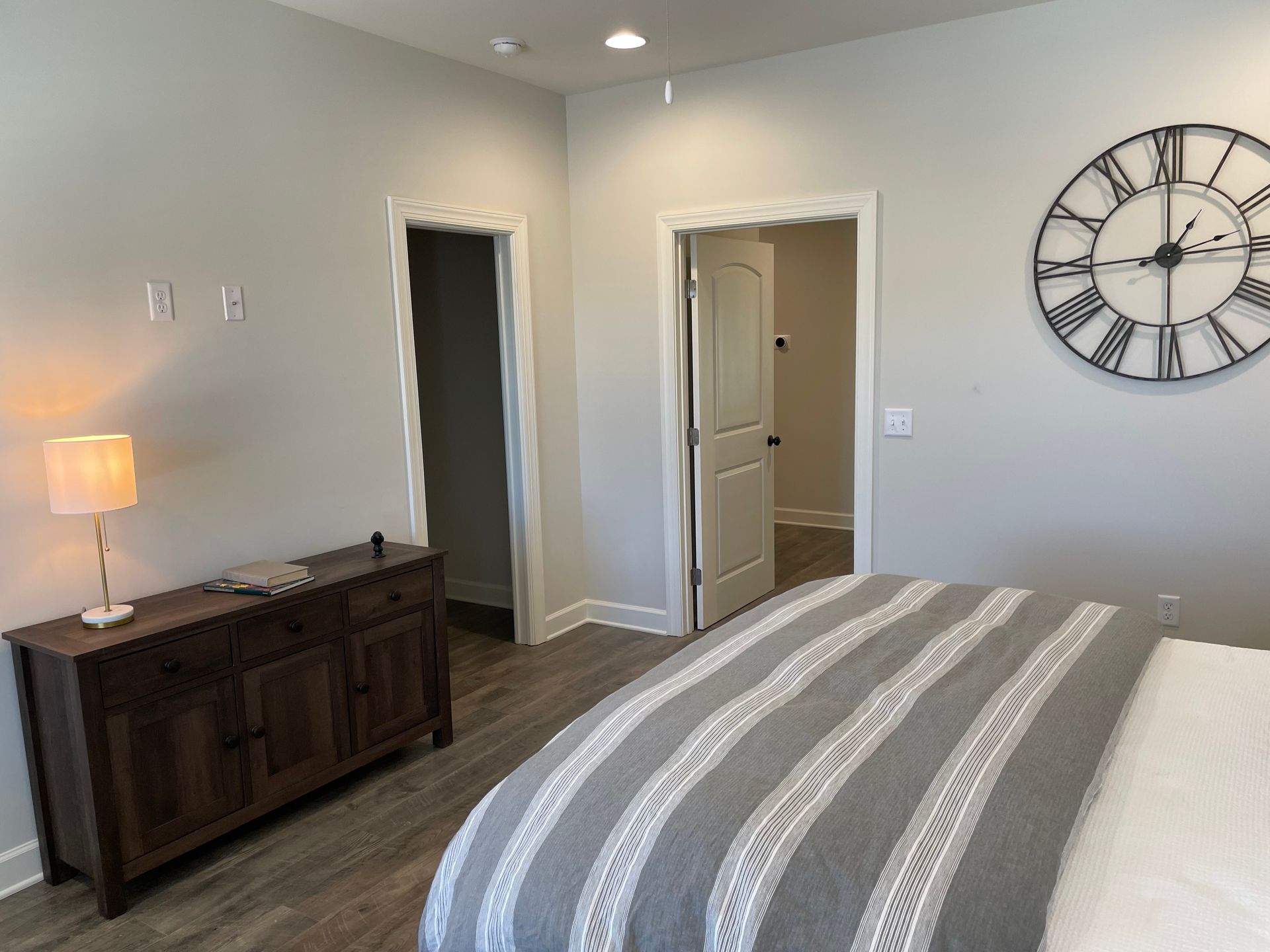 Bedroom with dresser, large clock, bed with gray striped blanket, and doorways.