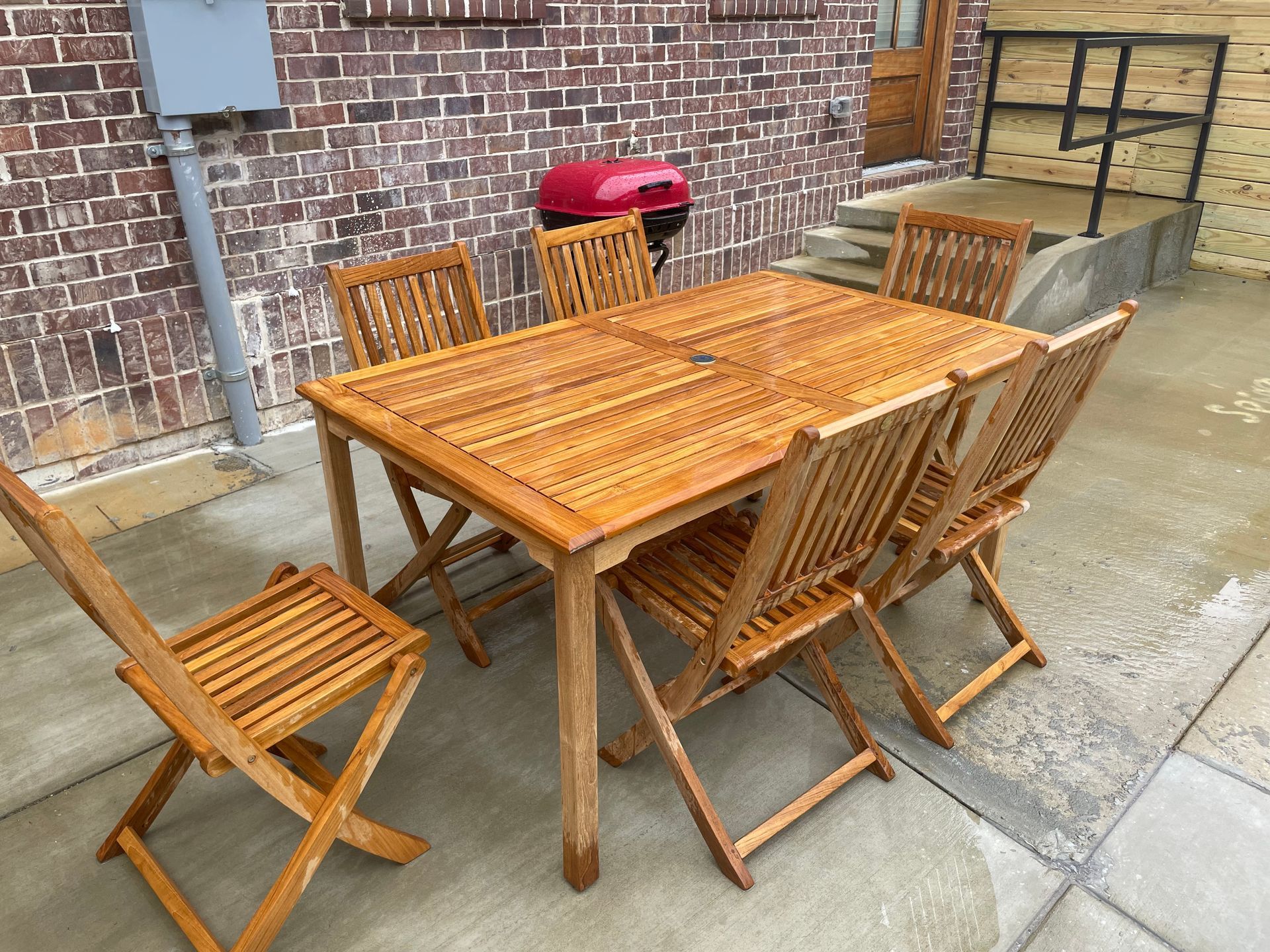 Wooden outdoor dining set on a patio, with a red grill and brick wall in the background.
