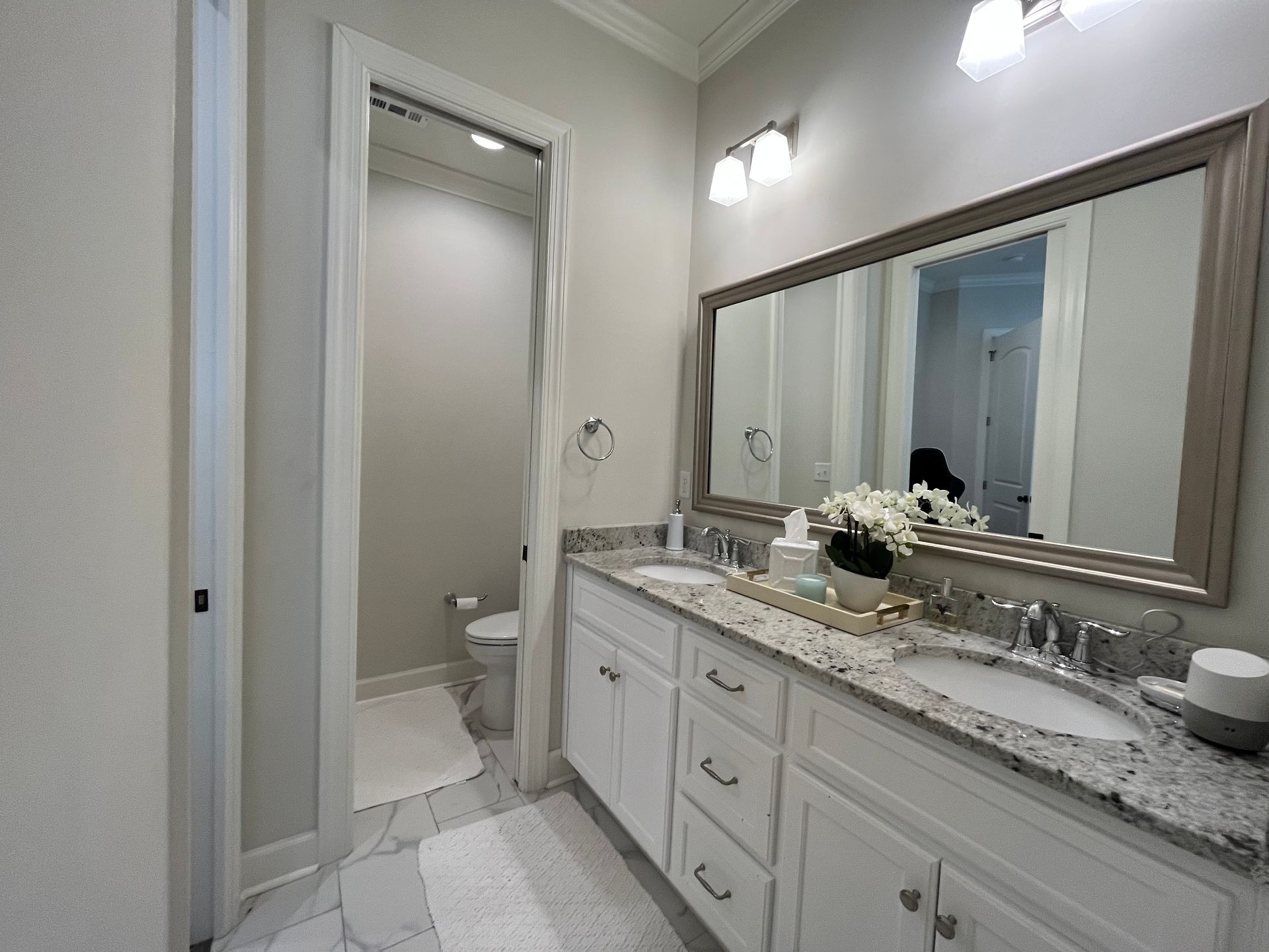 Bathroom with a white double vanity, granite countertop, and large mirror. A toilet is visible in the adjacent room.