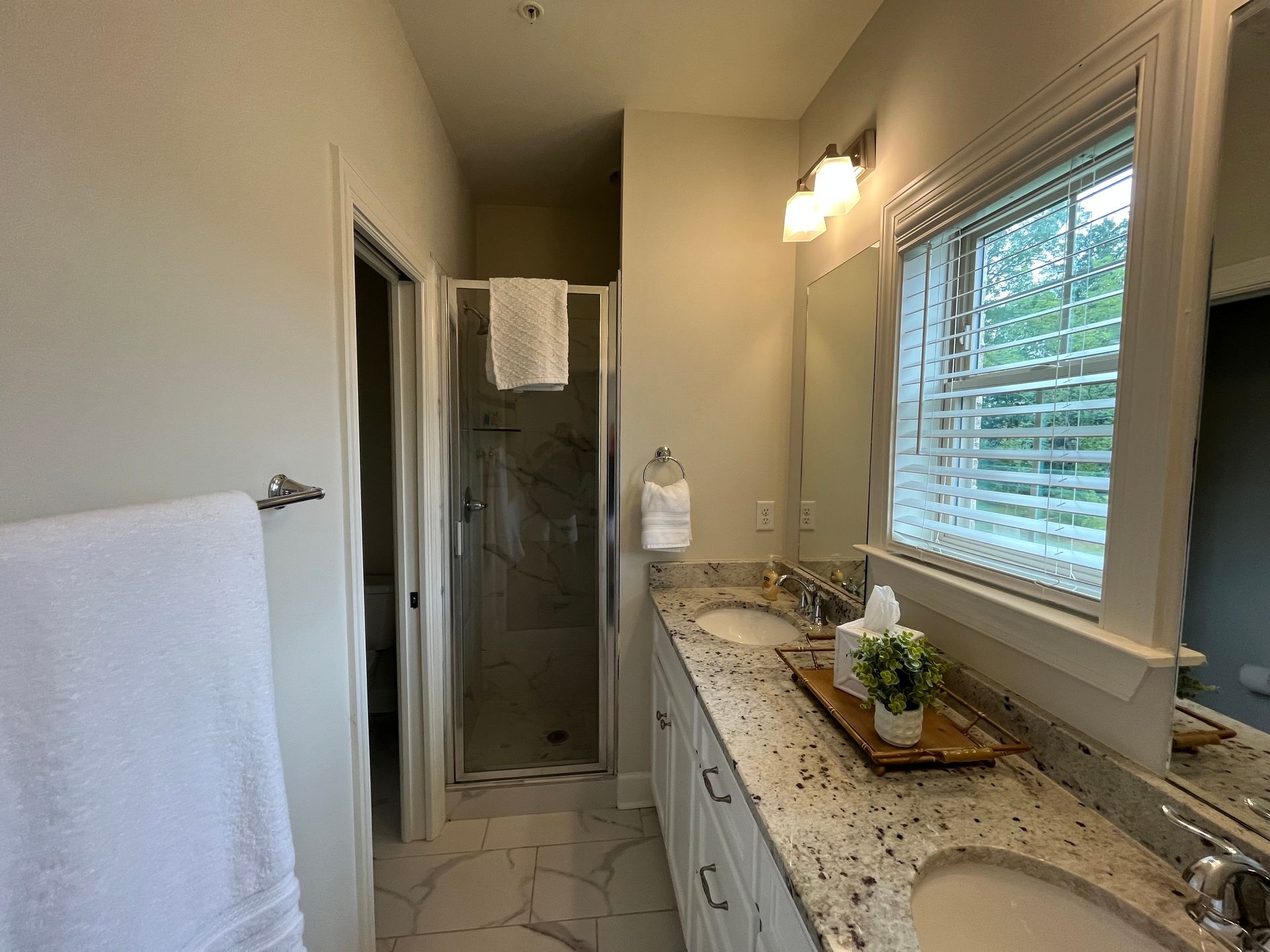 Bathroom with a granite countertop, double sinks, a shower, a window with blinds, and a large mirror.