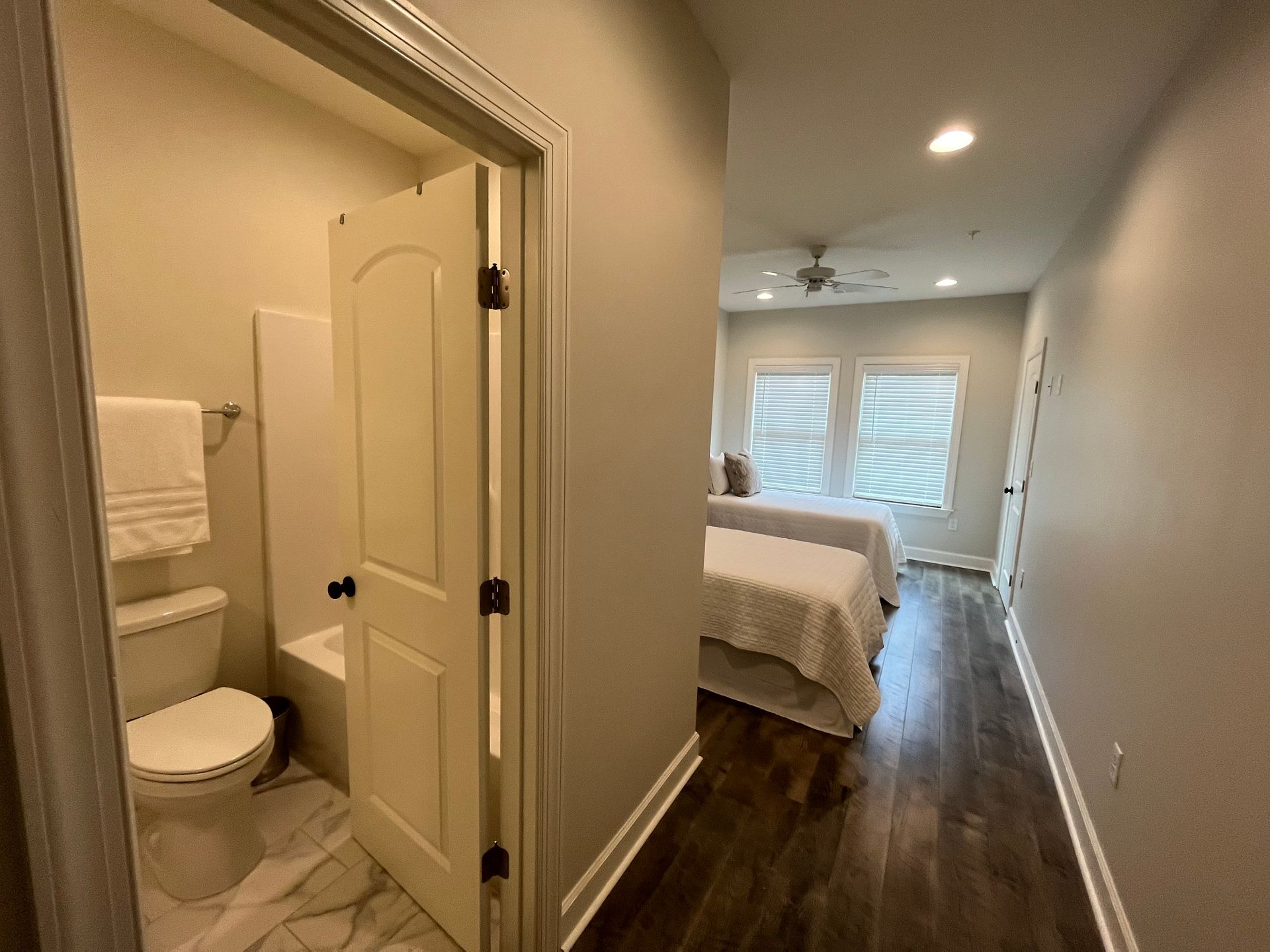 Hallway with bathroom door open, leading to a bedroom with two windows and a bed.
