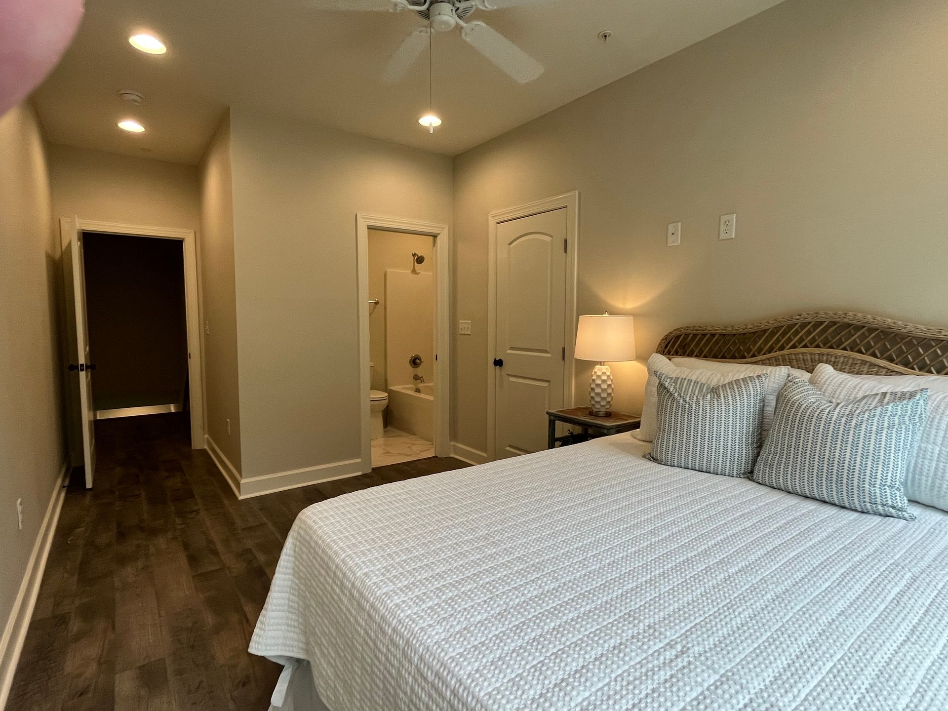 Bedroom with bed, bathroom, hallway, and ceiling fan. Neutral colors, wooden floor.