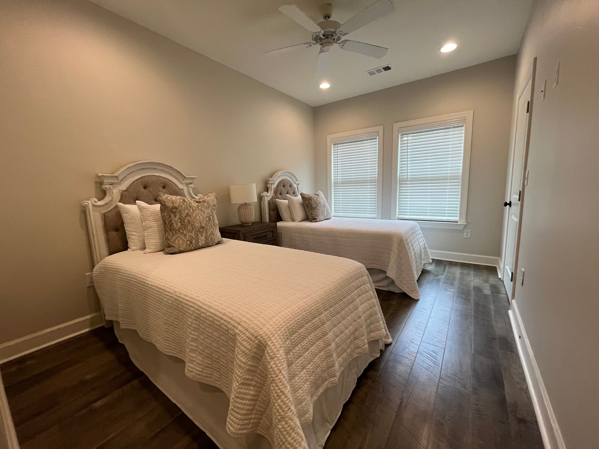 Two beds in a room, beige bedding, dark wood floor, two windows with blinds, neutral walls.