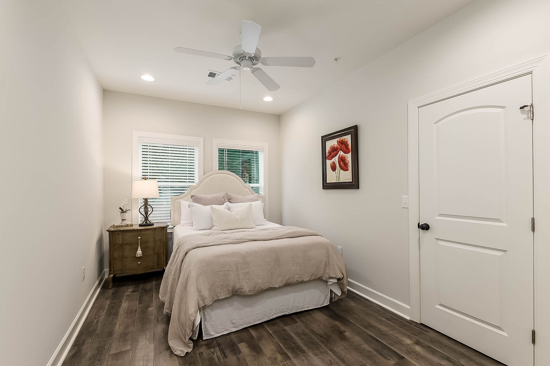 Bedroom with bed, nightstand, and window. White walls, wooden floor, artwork, and door.