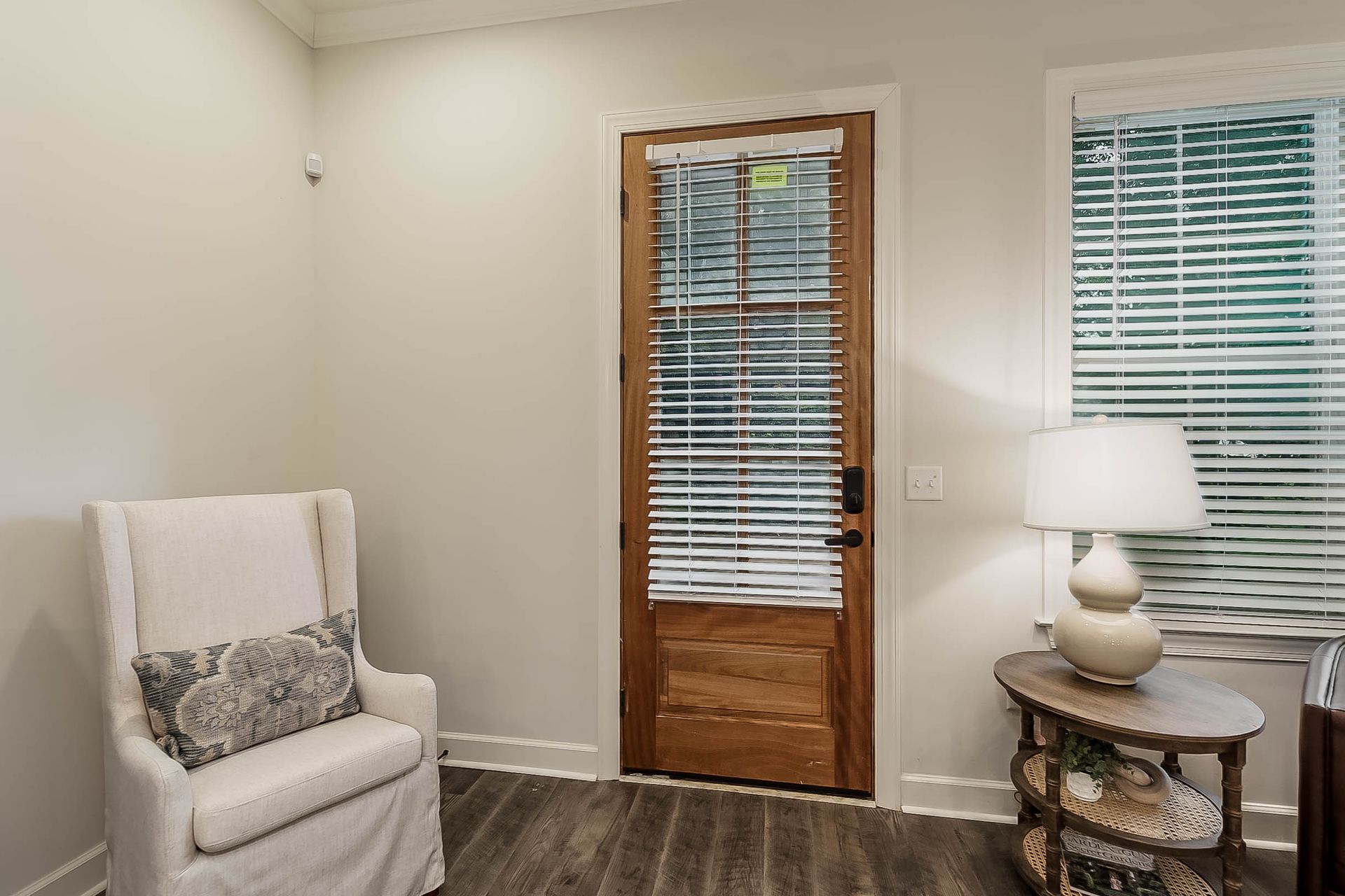 Living room with a white armchair, wooden door, oval side table, and window with blinds.