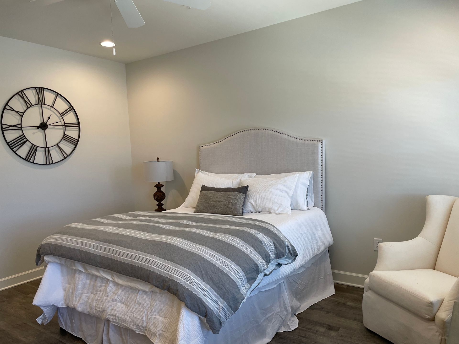 Bedroom with a bed, a white armchair, and a large wall clock. Gray and white bedding.
