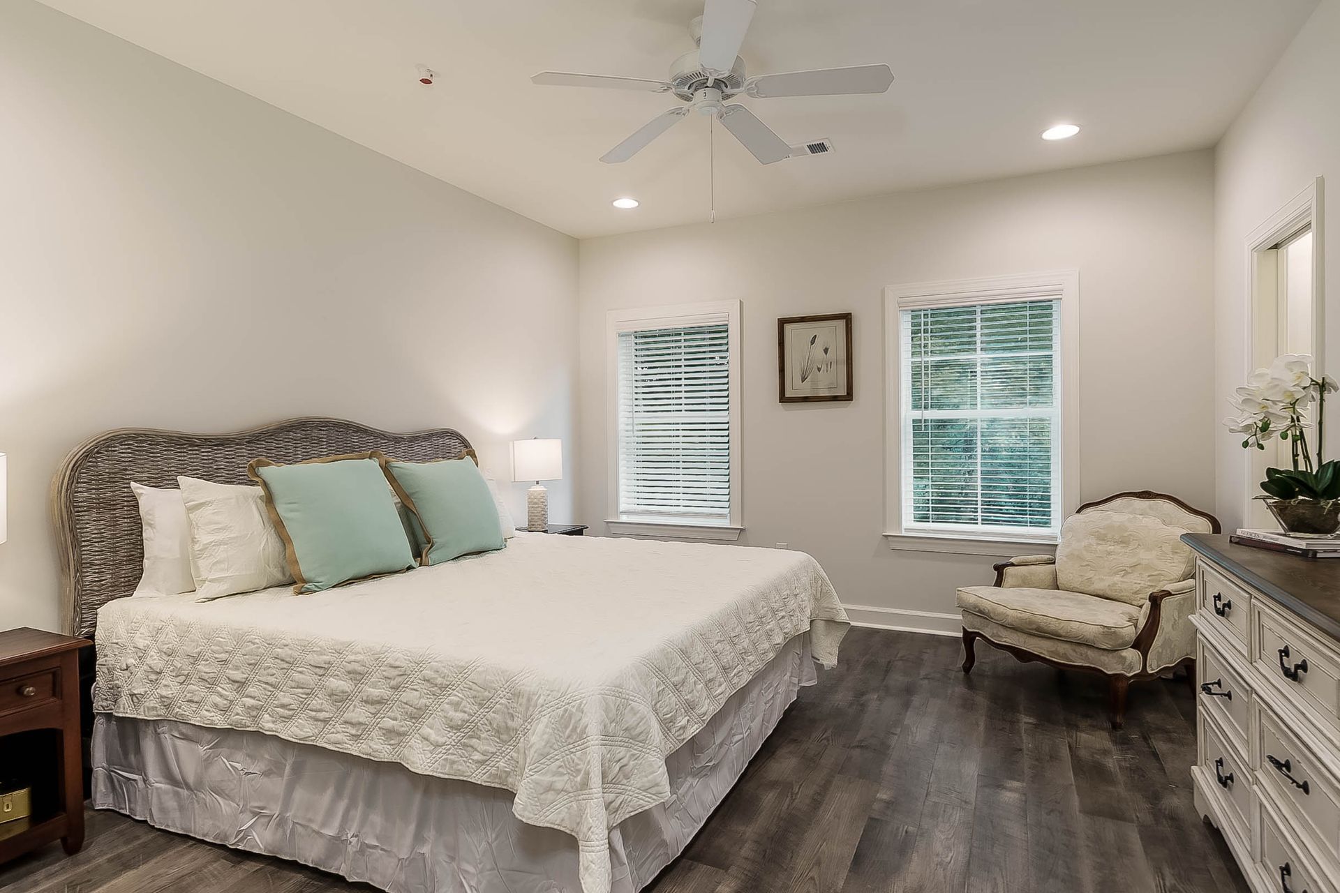 Bedroom with white walls, a bed with a woven headboard, two windows, wood flooring, and a patterned armchair.