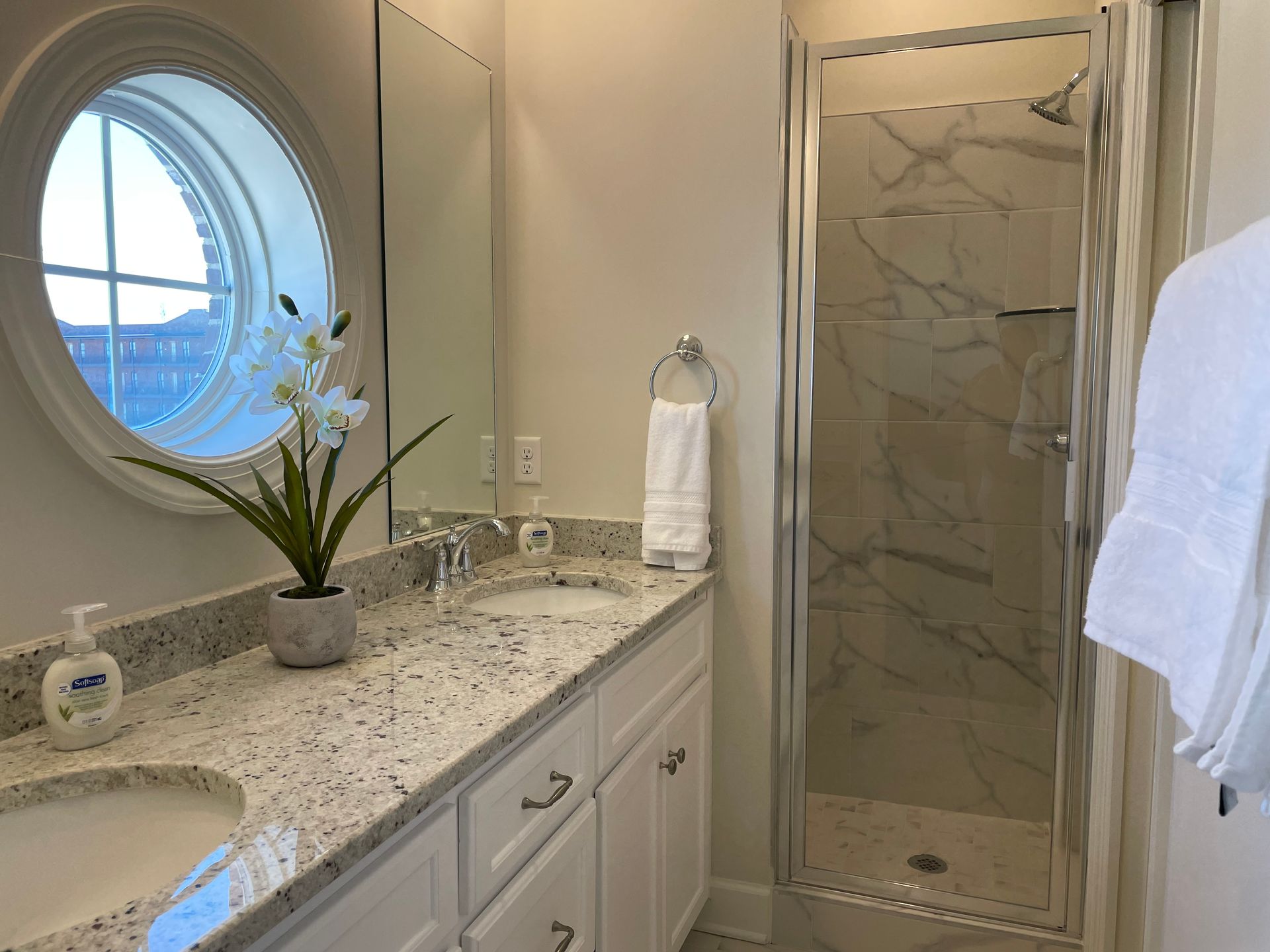 Bathroom with oval window, double vanity, and glass shower. White cabinets, marble-look shower tile.