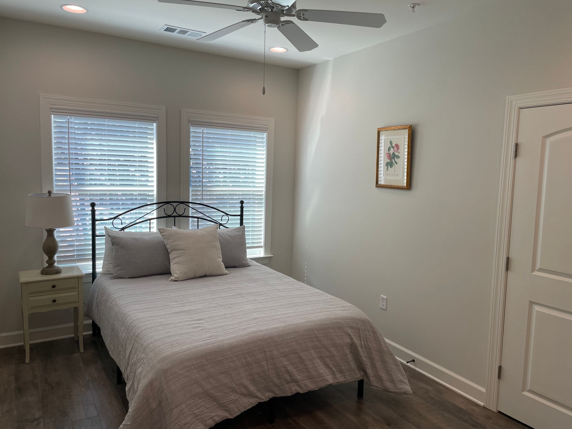 Bedroom with a bed, two windows with blinds, a nightstand, and a framed picture on the wall.