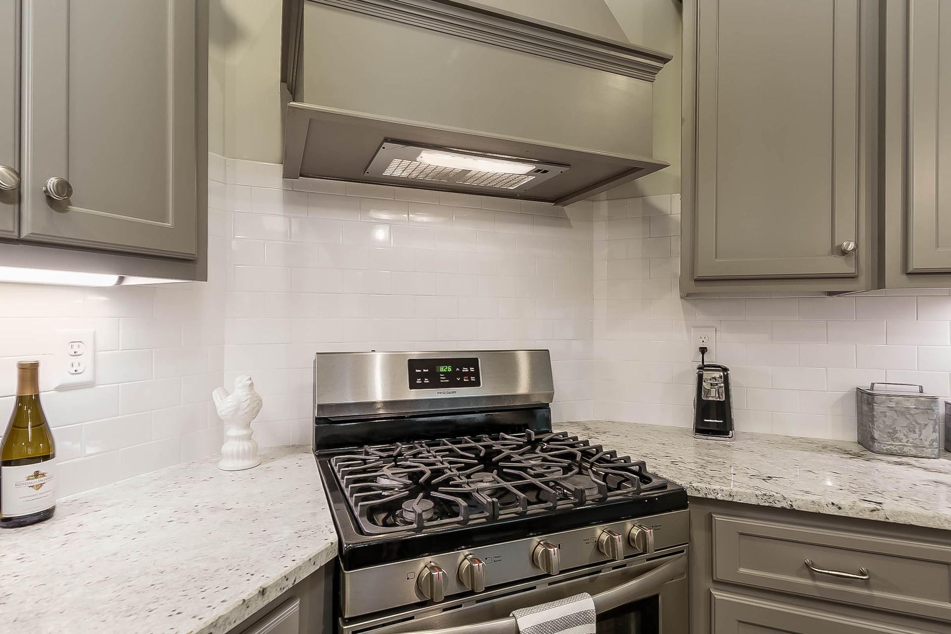 Kitchen corner with stainless steel stove, gray cabinets, white backsplash, and a range hood.