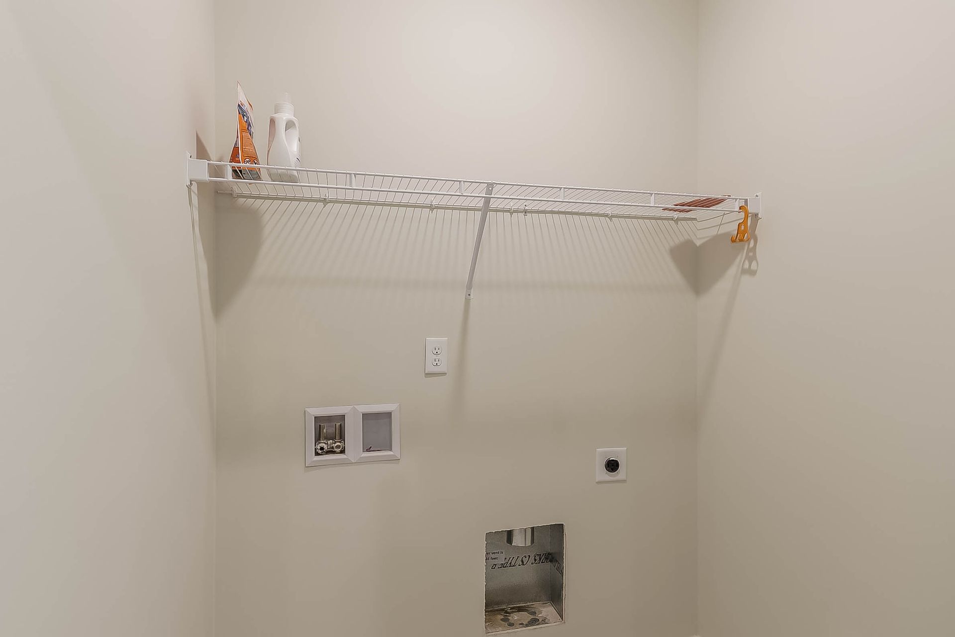 Laundry room with white wire shelf holding cleaning products. Wall has outlets and hookups.