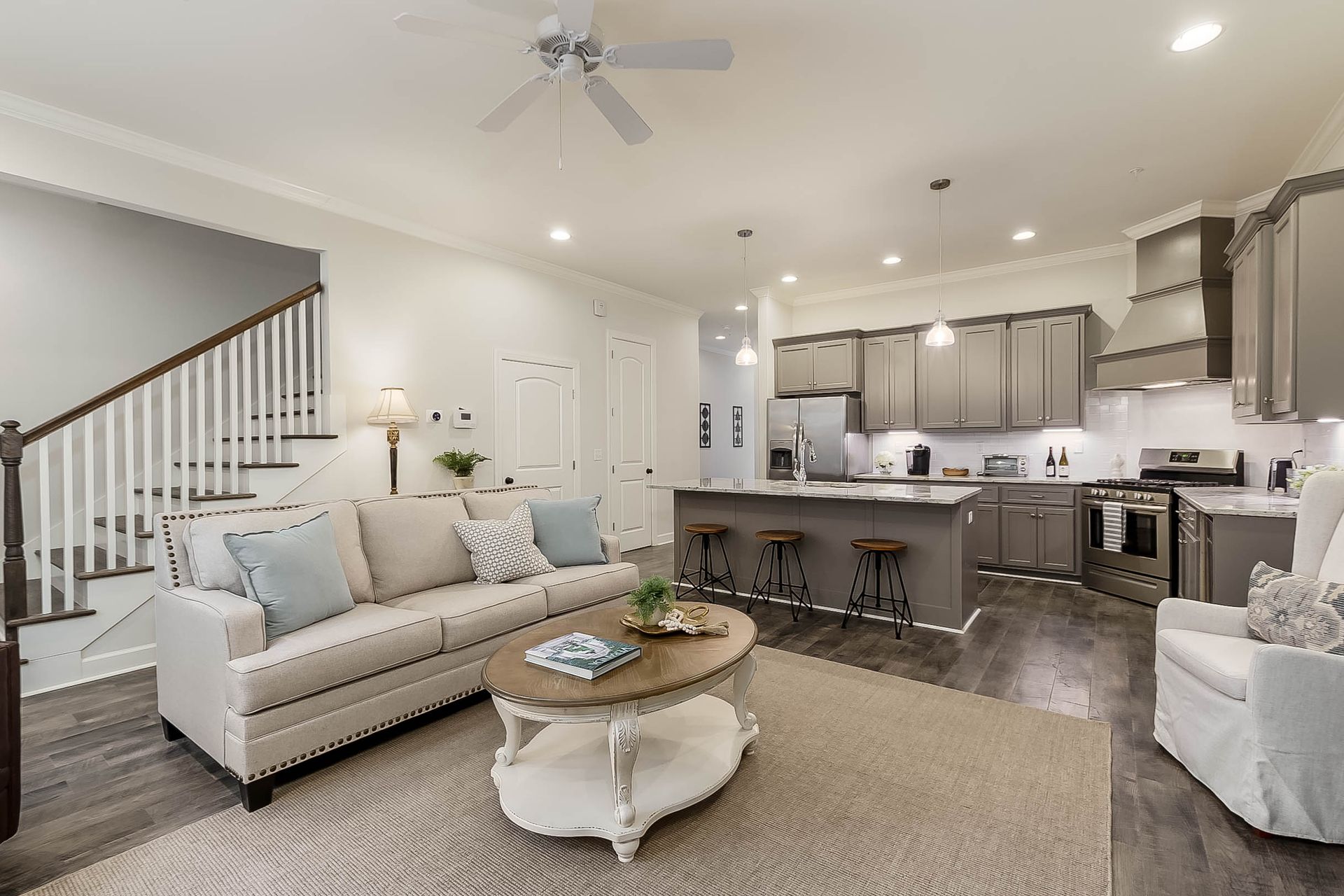 Open-concept living space with kitchen and living room; gray cabinets, beige couch, and wooden staircase.