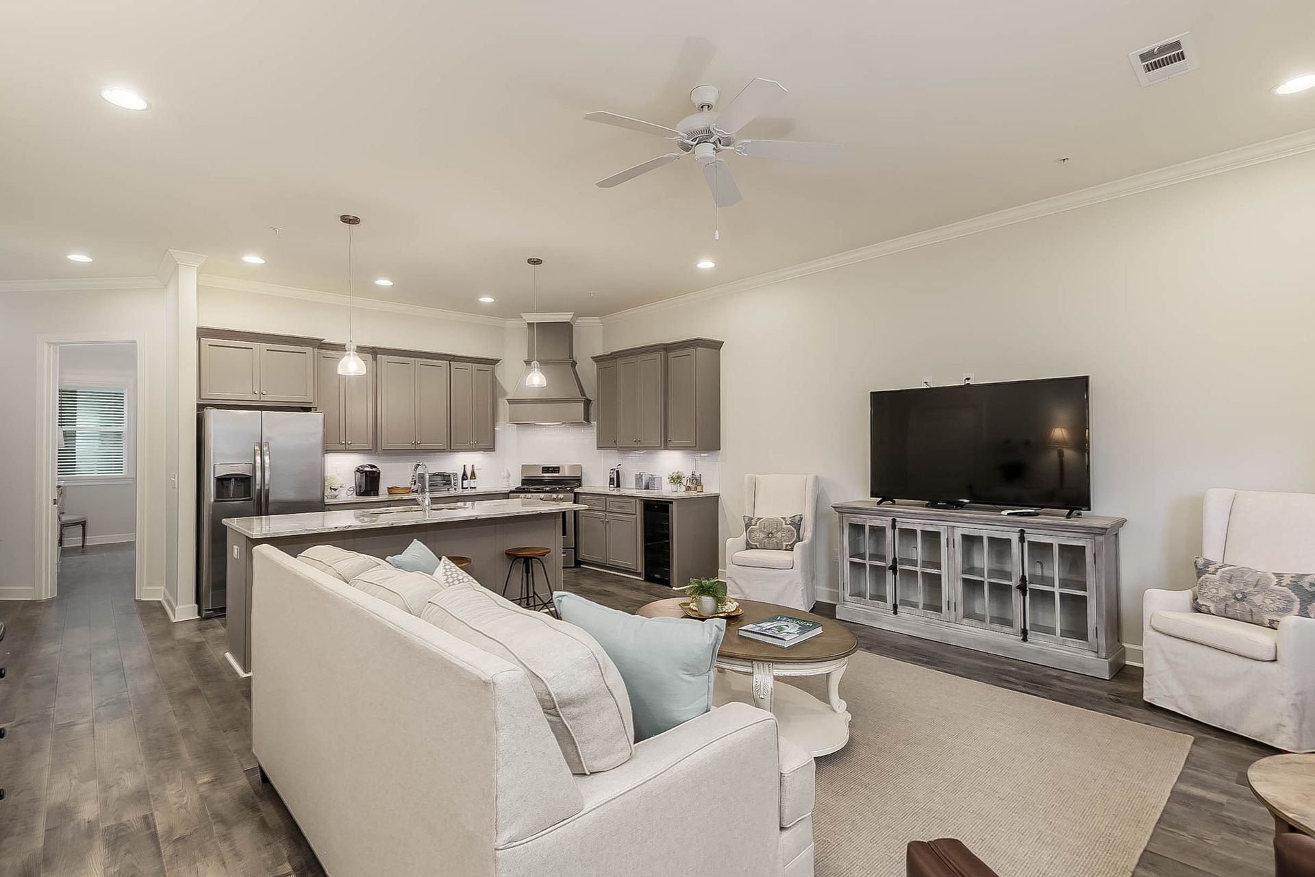 Living room with kitchen. Gray cabinetry, stainless steel appliances, white sofa, TV, and two armchairs.