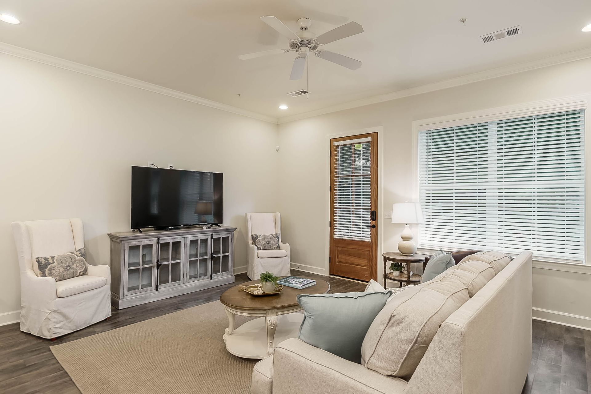 Living room with light-colored walls and furniture. Includes a TV, sofa, chairs, rug, coffee table, and window.