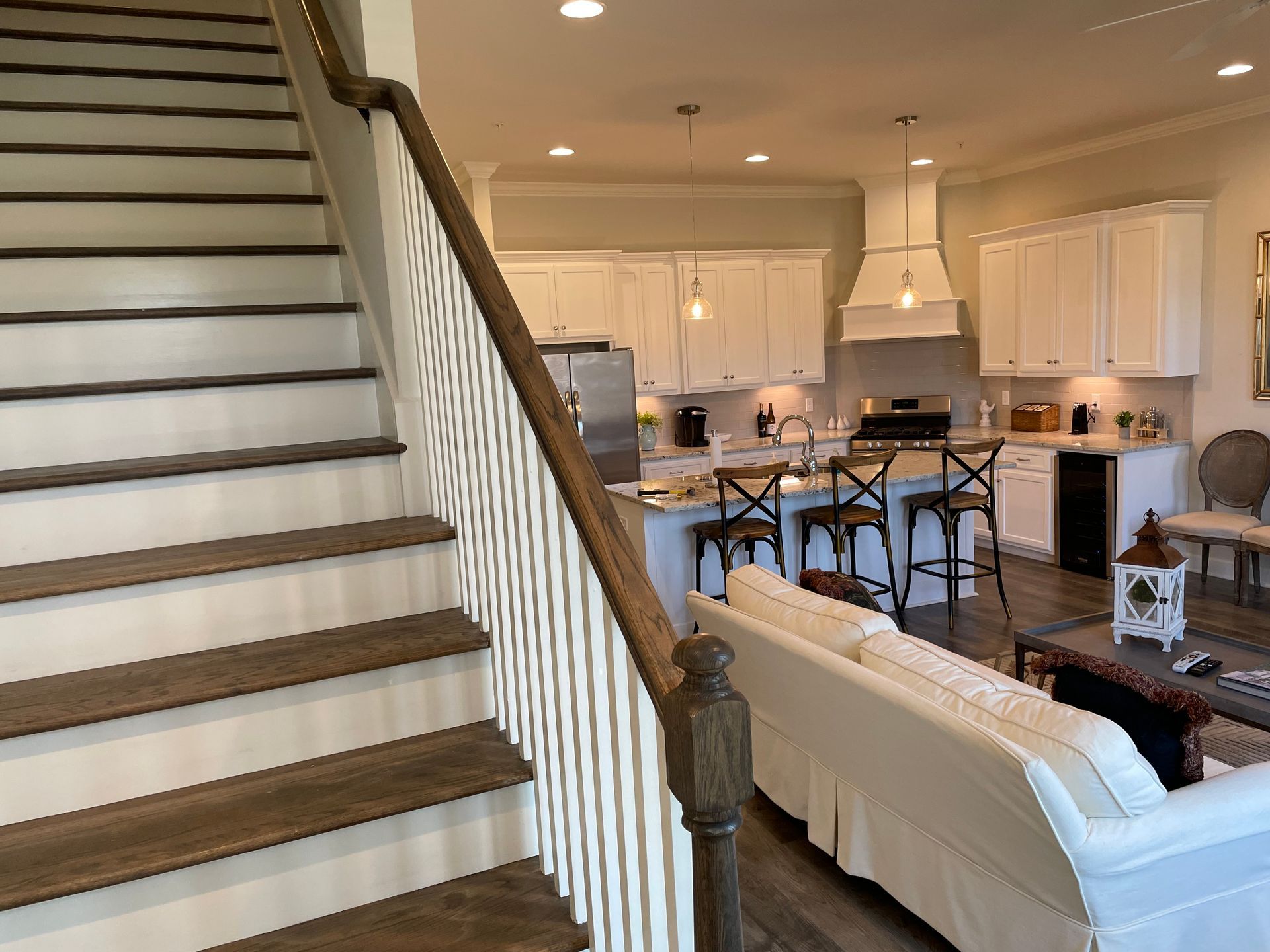 Wooden staircase leads to a light-filled kitchen with white cabinets, island, and seating area.