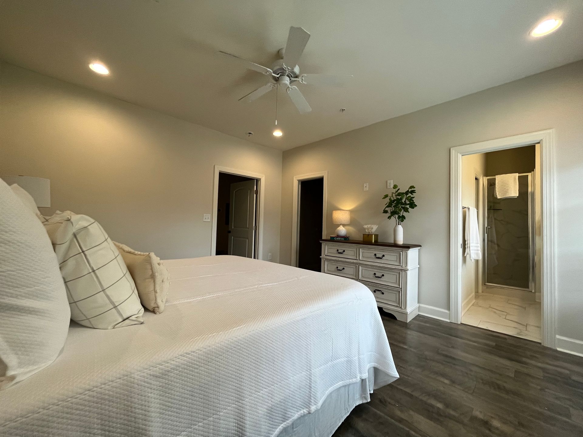 Spacious bedroom with white bedding, dresser, and three doorways. Gray walls, wood-look floor, and ceiling fan.
