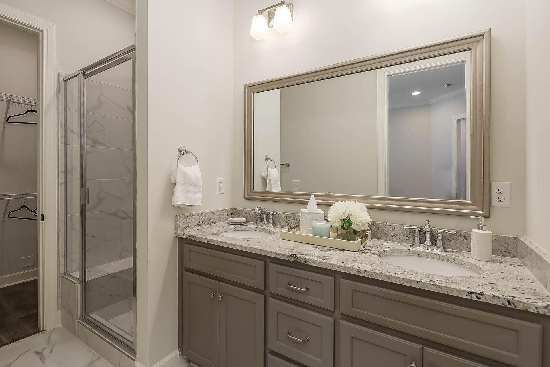 Bathroom with double sinks, gray cabinets, marble countertops, large mirror, and glass shower.