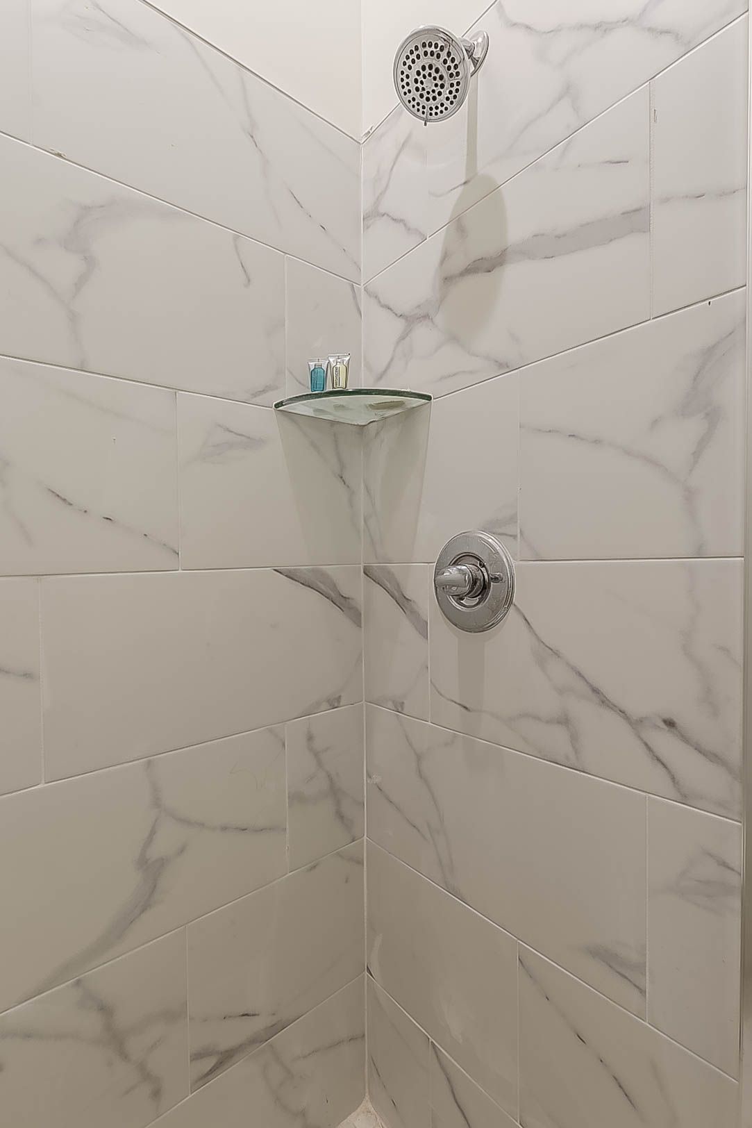 White tiled shower with glass shelf and chrome fixtures.