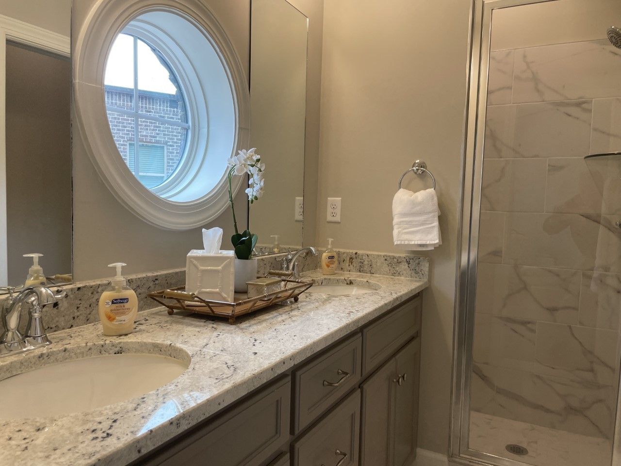 Bathroom with double sinks, gray cabinets, granite countertop, oval window, and a glass shower.