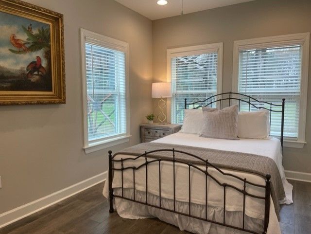 Bedroom with wrought iron bed, three windows, painting, and nightstand with lamp.