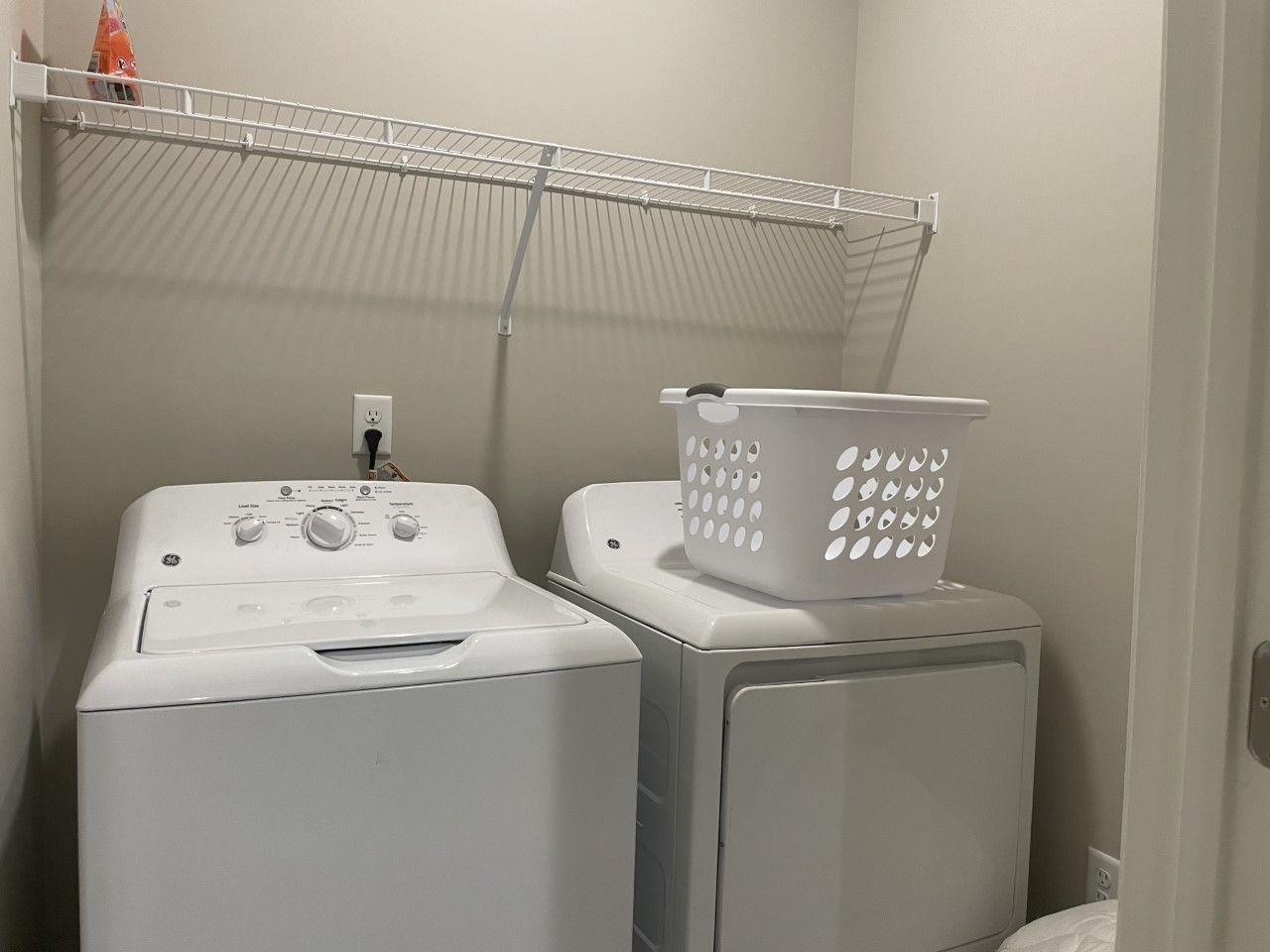 White washer and dryer in a laundry room with a wire shelf above. A laundry basket sits on the dryer.