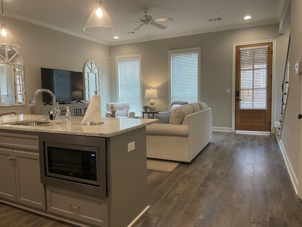 Open-concept living space with kitchen island, sofa, TV, and wooden door; gray walls, wood floors.