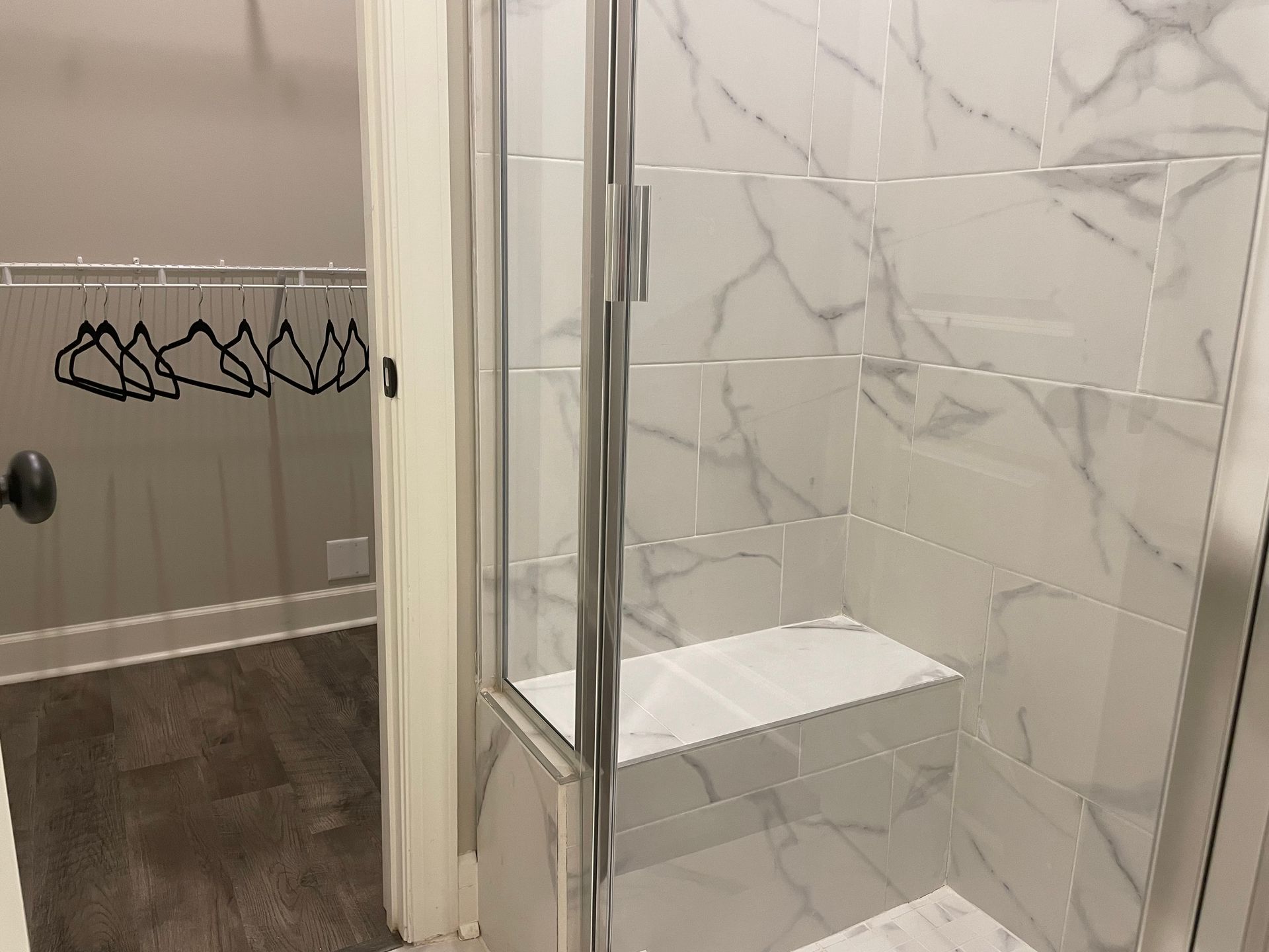 Shower with white marble-patterned tile, open glass door, and built-in bench. Closet visible in the background.