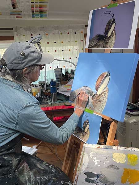 Artist wearing a cap painting a detailed portrait of a bird on a canvas, with a reference photo displayed above.
