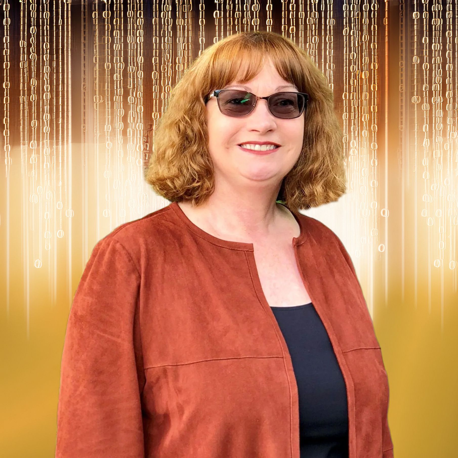 Woman with reddish hair and glasses smiles, wearing a rust-colored jacket over a black top, outdoors by green foliage.