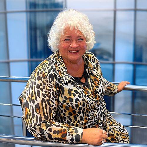 Woman in leopard-print shirt smiles at the camera, leaning on a railing. Blue glass building in background.