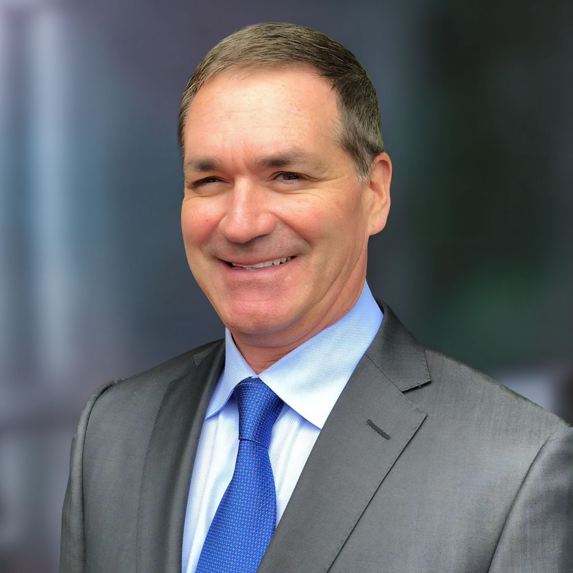 Man in suit and tie smiling against a blurred office backdrop.