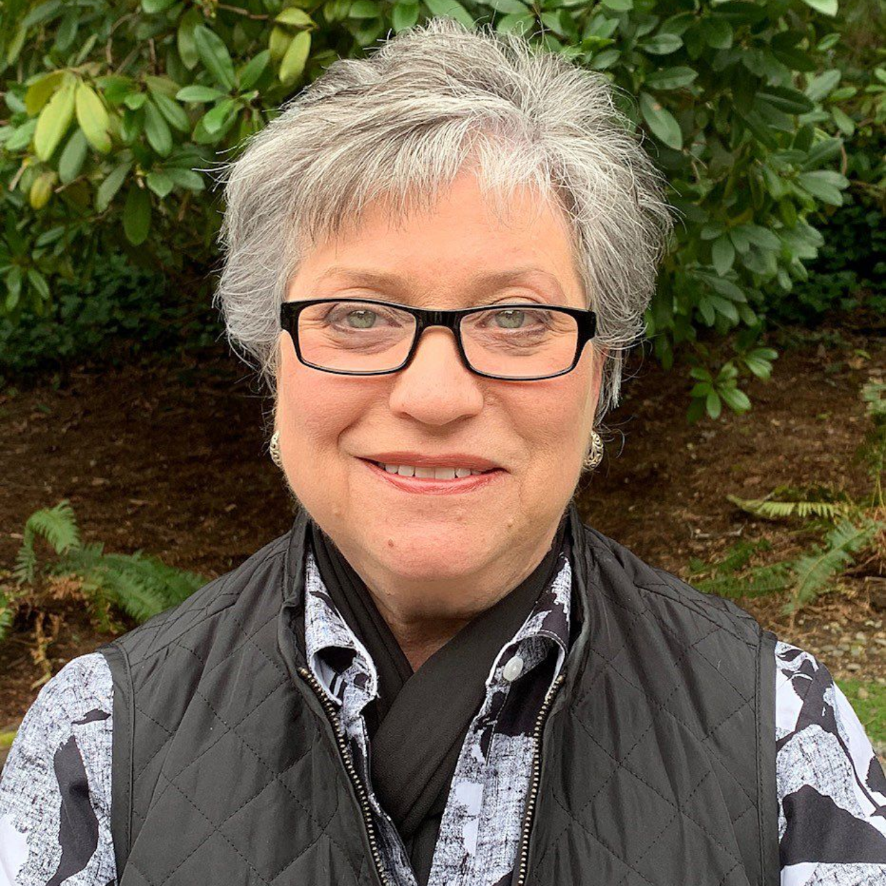 Woman with gray hair and glasses smiling outdoors, wearing a vest and patterned shirt.