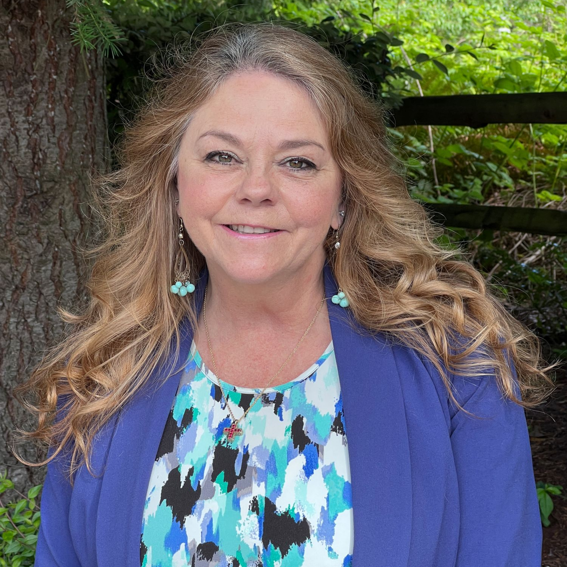 Woman with wavy blonde hair, smiling, wearing a blue blazer over a colorful top, outdoors.