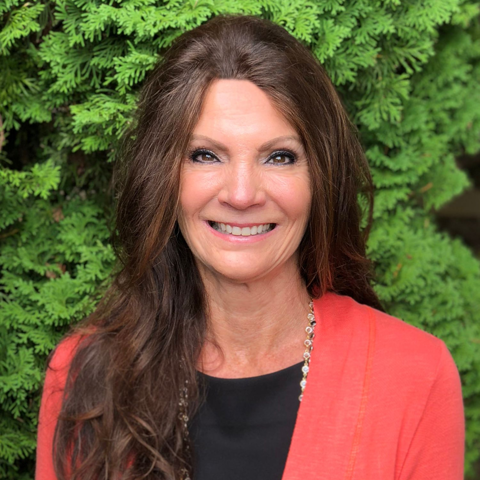 Woman with long brown hair smiles, wearing an orange cardigan over a black top, with green foliage background.