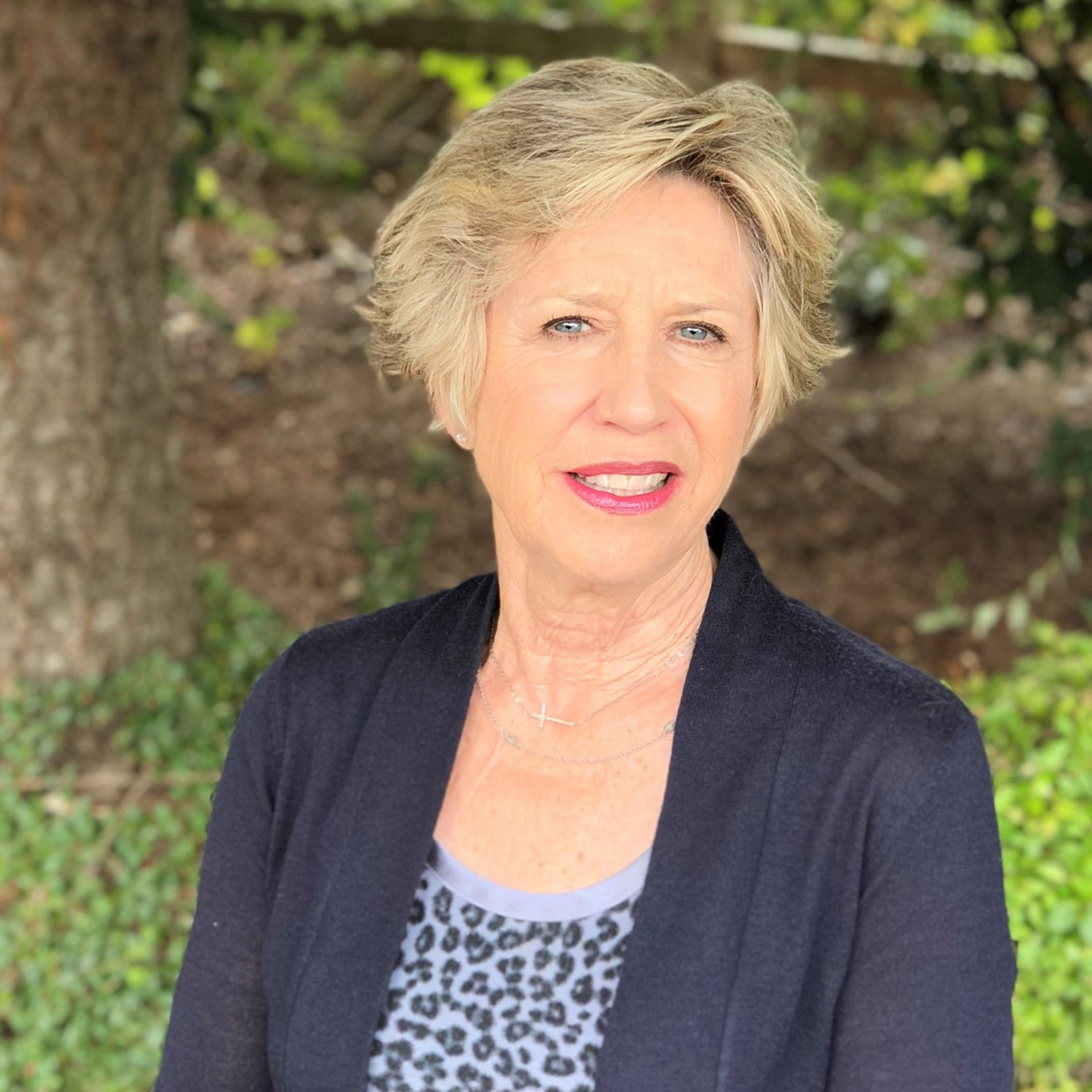 Woman with short blonde hair, smiling, wearing a dark blue cardigan over a patterned top, outdoors.