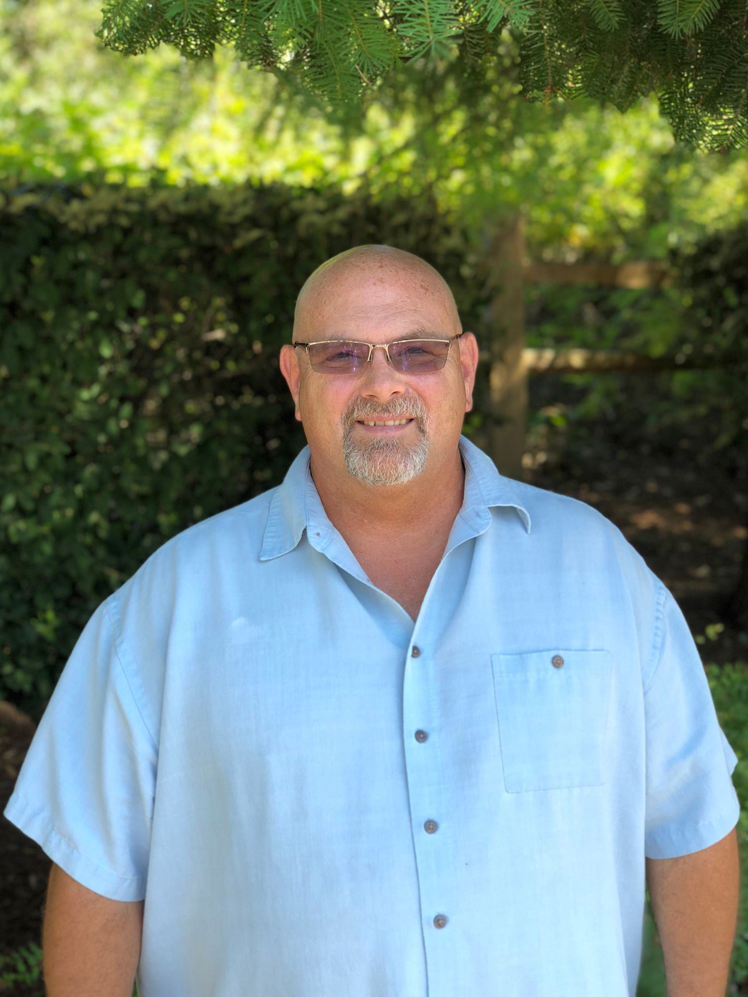 Man with a shaved head and glasses, smiling, wearing a light blue button-down shirt outdoors in front of greenery.