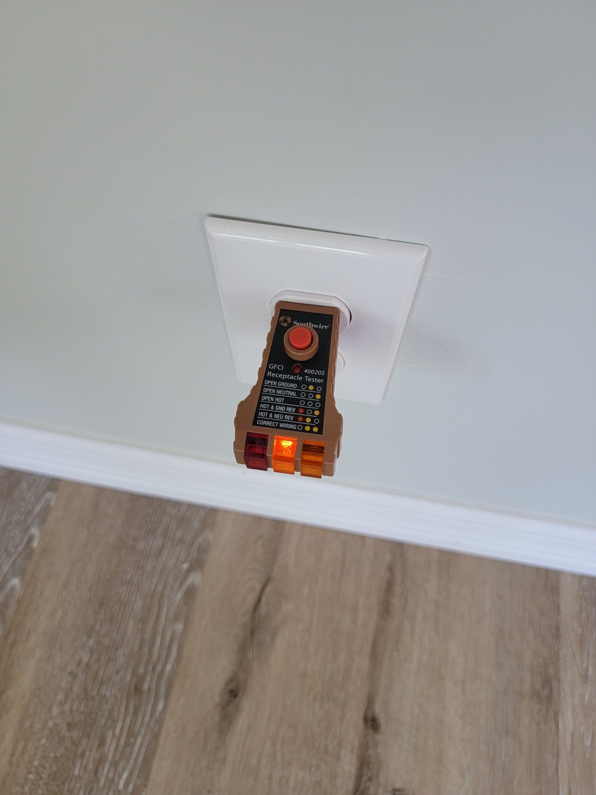 Electrical outlet tester plugged into a white outlet on a wall. Orange light illuminated.