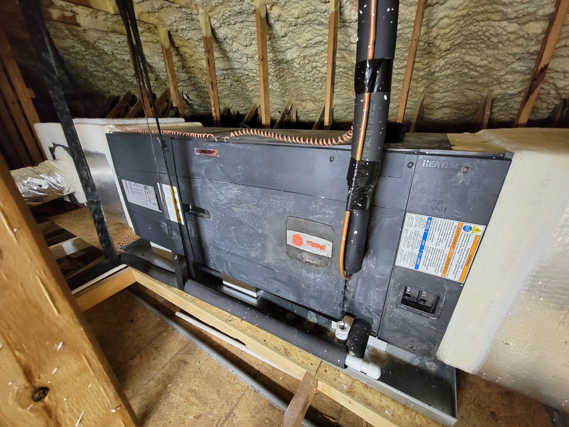 HVAC unit in an attic. Gray metal unit with black insulation. Wooden beams and insulation visible.