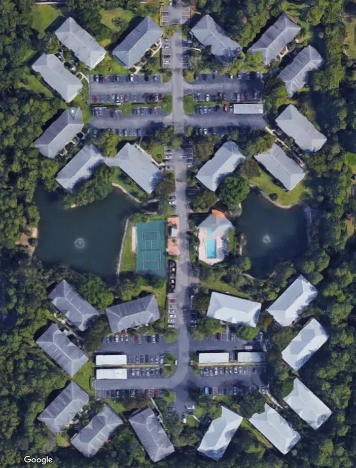 Aerial view of apartment complex with several buildings, two ponds, a pool, and a tennis court, surrounded by trees.