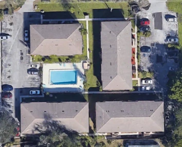 Aerial view of apartment complex with four buildings, swimming pool, and parking. Brown roofs, blue pool.