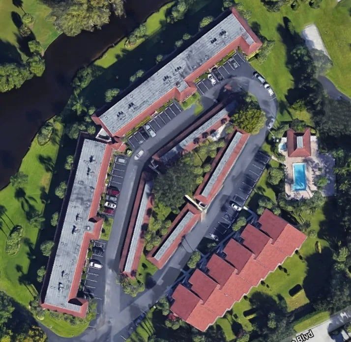 Aerial view of a condominium complex with red tile roofs, a pool, and parking. Green grass surrounds the buildings.