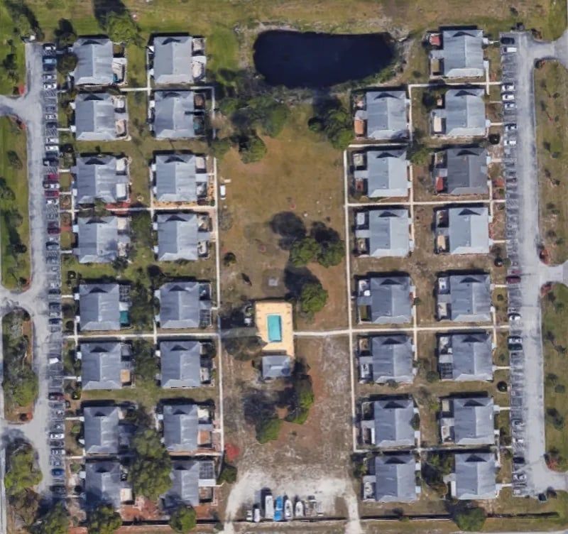 Aerial view of a rectangular apartment complex with a pool, pond, and parking areas surrounded by vegetation.