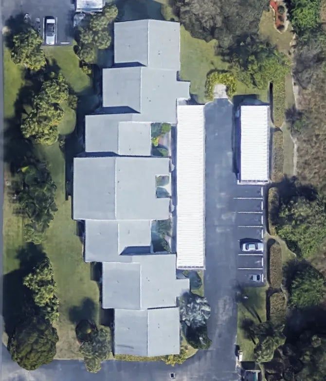 Aerial view of a complex with multiple gray roof sections, surrounded by green grass and trees. Parking lot visible.